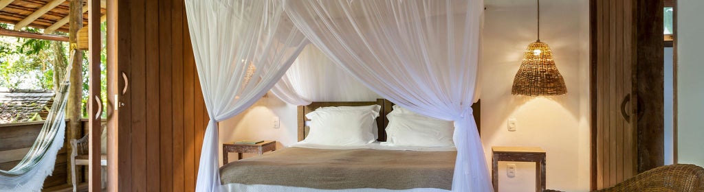 Elegant Brazilian hotel suite with wooden floors, minimalist décor, king-sized bed, and large windows overlooking lush tropical landscape in Trancoso