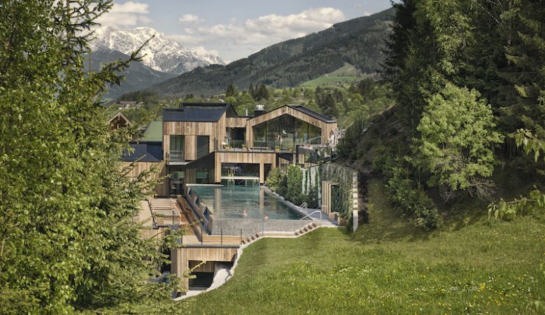Modern alpine spa hotel with infinity pool overlooking snow-capped mountains and forest, illuminated by warm evening light