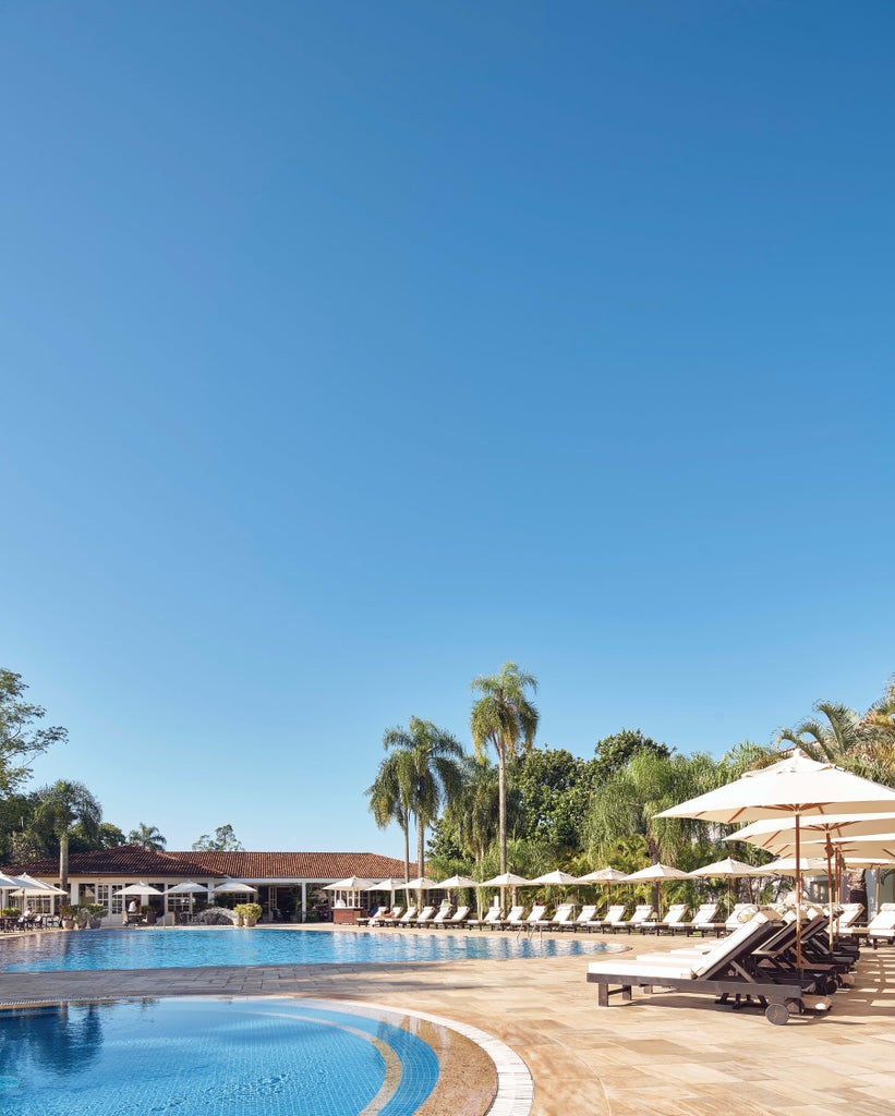 Pastel-pink Portuguese colonial hotel with white trim, surrounded by lush gardens and palm trees against a backdrop of Iguazu Falls