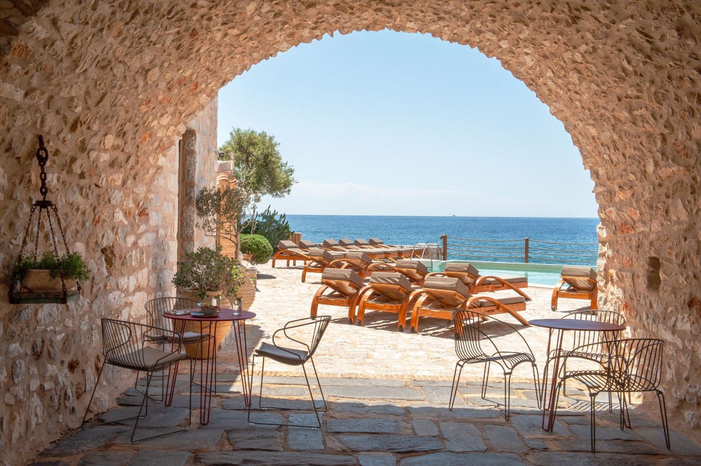 Luxurious stone seaside hotel with blue-green Aegean waters, traditional Greek architectural details, and sun-drenched stone terraces overlooking coastal scenery