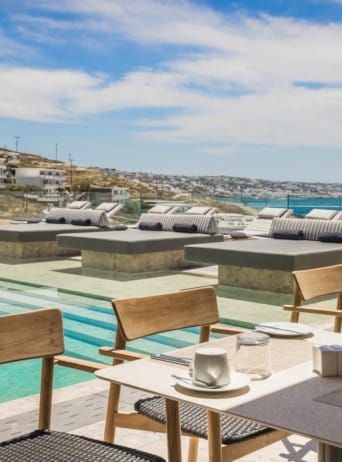 Luxurious white-washed hotel terrace overlooking azure Aegean Sea, featuring elegant minimalist design with sun loungers and panoramic Mykonos coastline