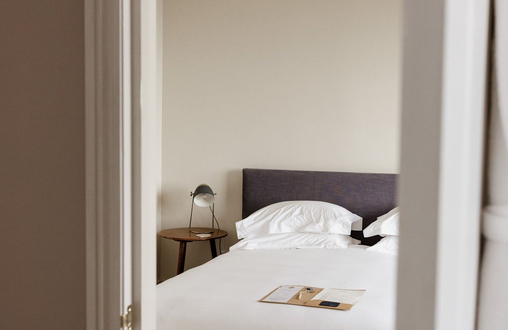 Elegant boutique hotel bedroom with crisp white linens, plush throw pillows, wooden floors, and soft natural lighting in a contemporary UK design