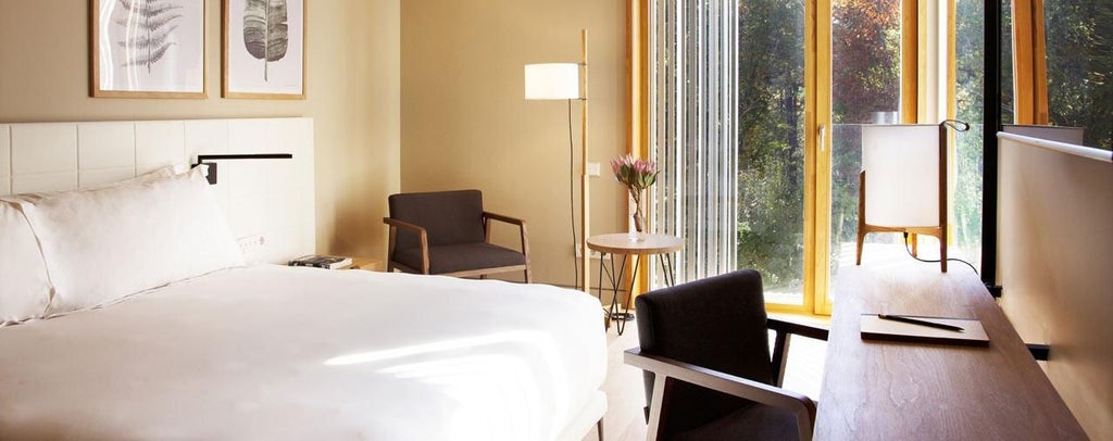 Elegant superior hotel room with minimalist design, plush white bedding, modern wooden furnishings, and expansive windows overlooking Spanish landscape at sunset