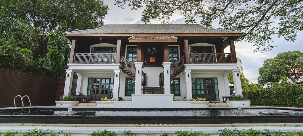 Traditional Thai boutique hotel with teak wood balconies, white colonial-style architecture, and lush tropical gardens beside a river