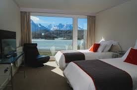 Modern rustic hotel with wooden paneling nestled against snowy mountains, reflected in Grey Lake amid Torres del Paine's wilderness