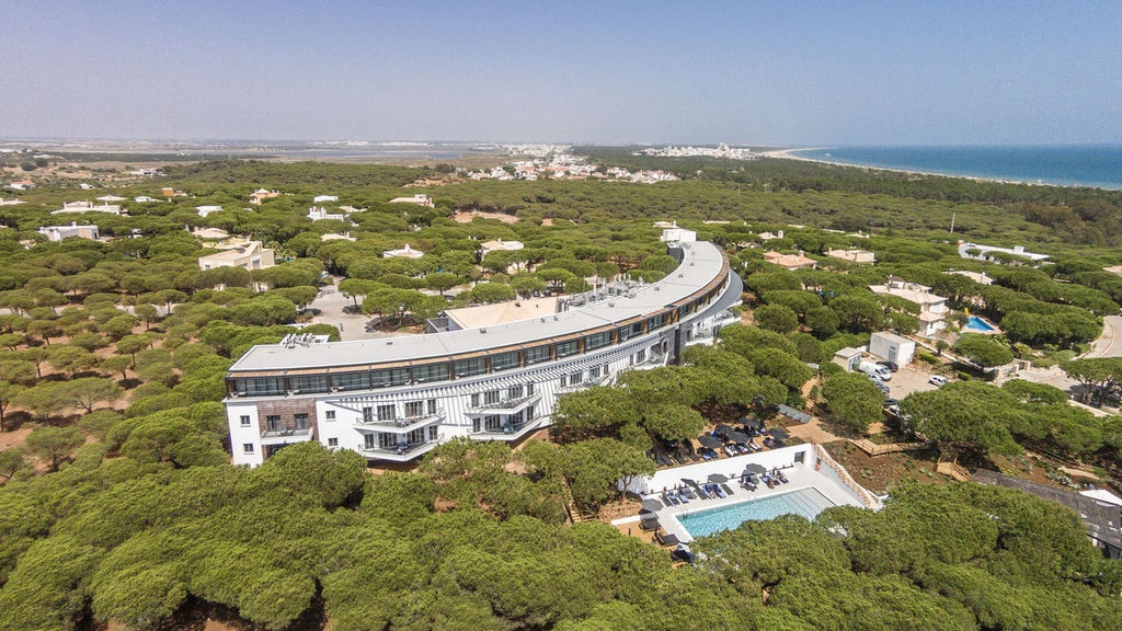 Sleek white hotel building surrounded by lush pine forest, featuring modern architecture and glass balconies overlooking Mediterranean sea