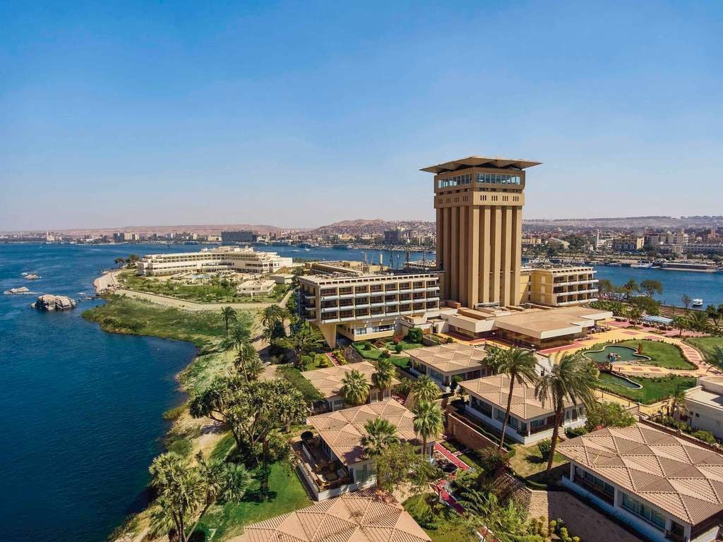 Luxurious historic Sofitel Legend hotel nestled along the Nile River, featuring elegant Nubian-style architecture with golden stone walls and lush tropical gardens.