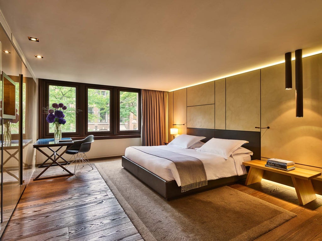 Elegant modern deluxe hotel room with warm wood tones, plush furnishings, and large windows overlooking Amsterdam's historic architectural landscape